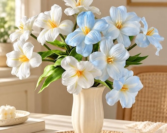 May include: A bouquet of artificial flowers in a cream-colored fluted vase. The arrangement features white and light blue petals with yellow centers, accented by green leaves. The flowers are displayed in a tall, slender ceramic vessel.