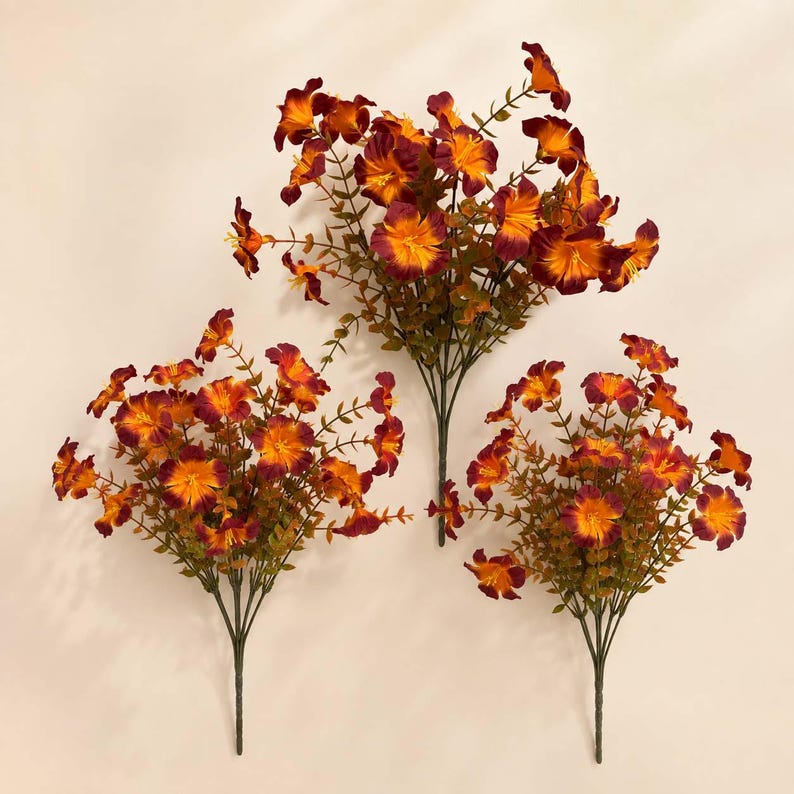 May include: Three artificial flower bushes featuring vibrant orange and deep burgundy petals. Each stem consists of multiple small blossoms and green foliage arranged in a cluster, creating a full, rounded appearance for each individual floral spray.