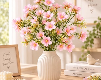 May include: A bouquet of artificial light pink flowers with yellow centers and green stems arranged in a white, vertically ribbed ceramic vase. The flowers feature five petals each and are displayed in a dense, rounded cluster with small green buds.