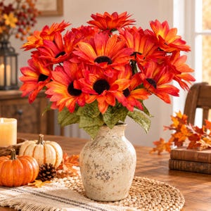 May include: A bouquet of vibrant orange and yellow artificial sunflowers with dark brown centers arranged in a speckled, off-white ceramic vase. The flowers feature layered petals and green leaves, creating a dense, rounded floral display.