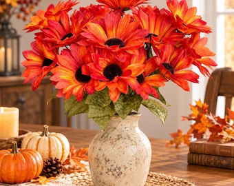 May include: A bouquet of vibrant orange and yellow artificial sunflowers with dark brown centers arranged in a speckled, off-white ceramic vase. The flowers feature layered petals and green leaves, creating a dense, rounded floral display.