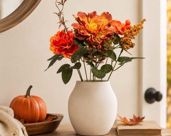 May include: A matte off-white ceramic vase containing a bouquet of artificial orange and rust-colored flowers, green leaves, and small yellow berries. The arrangement features large, layered petals and thin stems, creating a vibrant display.