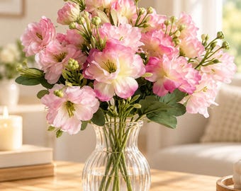 May include: A bouquet of artificial pink and white flowers with green stems and buds arranged in a clear, ribbed glass vase. The flowers feature layered petals with soft pink edges that fade into creamy white centers, creating a full, rounded floral display.