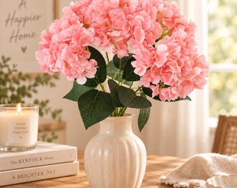 May include: A bouquet of artificial pink hydrangea flowers with green leaves arranged in a cream-colored, vertically ribbed ceramic vase. The flowers feature dense, rounded clusters of soft pink petals, providing a full and textured appearance.