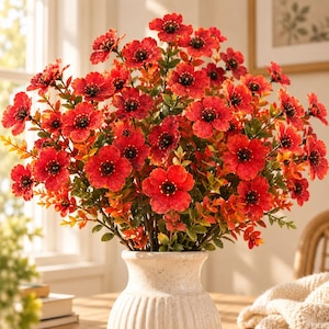 May include: A bouquet of vibrant red artificial flowers with dark centers and textured petals, arranged in a cream-colored ceramic vase with a ribbed base. The stems feature small green and orange leaves, creating a dense, rounded floral display.