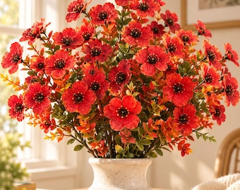 May include: A bouquet of vibrant red artificial flowers with dark centers and textured petals, arranged in a cream-colored ceramic vase with a ribbed base. The stems feature small green and orange leaves, creating a dense, rounded floral display.