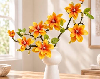 May include: A white ceramic vase with a rounded body and a flared base holds a branch of artificial flowers. The flowers feature vibrant orange and yellow petals with white centers, accompanied by small green leaves and fuzzy, light-colored buds.