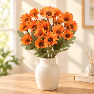 May include: A bouquet of artificial orange sunflowers with dark brown centers and green leaves arranged in a smooth, off-white ceramic vase. The flowers feature multiple layers of petals in varying shades of orange, creating a full, rounded floral display.