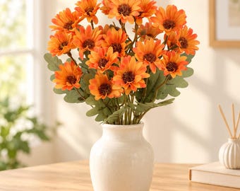 May include: A bouquet of artificial orange sunflowers with dark brown centers and green leaves arranged in a smooth, off-white ceramic vase. The flowers feature multiple layers of petals in varying shades of orange, creating a full, rounded floral display.