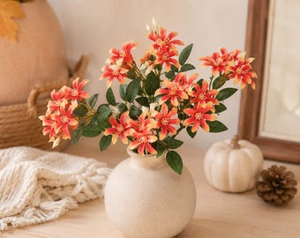 May include: A round, off-white ceramic vase holds a bouquet of artificial flowers with vibrant orange and yellow petals and dark green leaves. The vase has a smooth, matte texture and a narrow neck, contrasting with the bright, multi-colored floral arrangement.