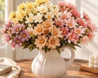 May include: A white ceramic pitcher vase with vertical fluted detailing holds a dense, multi-colored bouquet of artificial daisy-like flowers. The arrangement features petals in shades of yellow, cream, peach, pink, and light purple with green leaves.