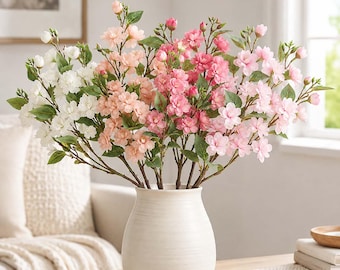 May include: A bouquet of artificial cherry blossom branches in a white ceramic vase. The branches feature clusters of flowers in shades of white, peach, light pink, and dark pink, interspersed with small green leaves and unopened flower buds.