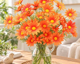 May include: A bouquet of vibrant orange daisies with bright yellow centers arranged in a clear glass pitcher vase with two side handles. The flowers feature multiple layers of petals and green stems visible through the glass, resting on a round woven mat.