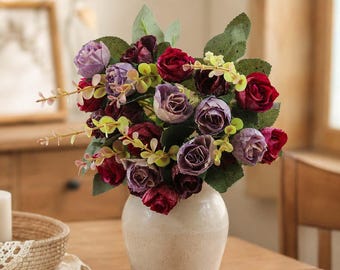 May include: A bouquet of artificial flowers featuring deep red and muted purple roses mixed with small green leaves and light yellow sprigs. The floral arrangement is displayed in a smooth, off-white ceramic vase with a rounded base and neck.