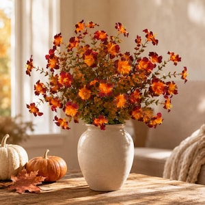 Puede incluir: Un ramo de flores artificiales con pétalos de color naranja vibrante y rojo intenso dispuesto en un jarrón de cerámica de color blanco roto. Los tallos tienen pequeñas hojas verdes y el arreglo se muestra junto a dos calabazas y una hoja.