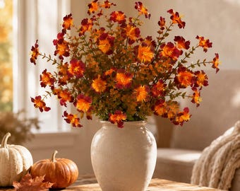 May include: A bouquet of artificial flowers with vibrant orange and deep red petals arranged in a smooth, off-white ceramic vase. The stems feature small green leaves, and the arrangement is displayed alongside two small pumpkins and a fallen leaf.