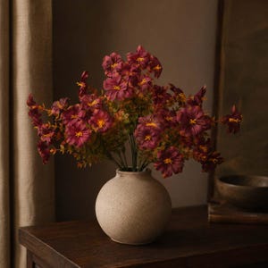 May include: A bouquet of deep burgundy flowers with yellow centers and green foliage arranged in a round, beige, textured ceramic vase. The flowers have multiple petals and are displayed on thin stems, creating a full, rounded floral arrangement.