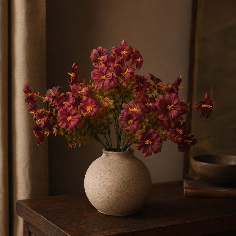 May include: A bouquet of deep burgundy flowers with yellow centers and green foliage arranged in a round, beige, textured ceramic vase. The flowers have multiple petals and are displayed on thin stems, creating a full, rounded floral arrangement.