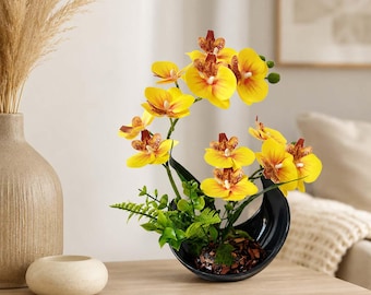 May include: A decorative arrangement of artificial yellow orchids with reddish-brown centers, featuring green leaves and fern-like foliage. The flowers are set in a curved, black crescent-shaped ceramic planter filled with dark brown decorative stones.