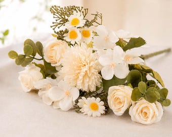 May include: A bouquet of artificial flowers featuring cream-colored roses, a central pale peach chrysanthemum, white daisies with yellow centers, and small white blossoms. The arrangement includes green eucalyptus-style leaves and stems.