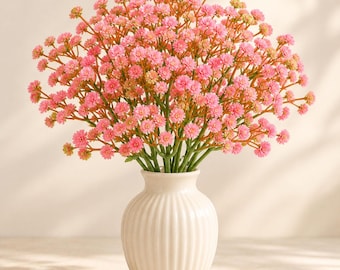 May include: A bouquet of artificial pink baby's breath flowers with small, clustered blossoms and green stems, arranged in a white, vertically ribbed ceramic vase with a rounded body and a narrow neck.
