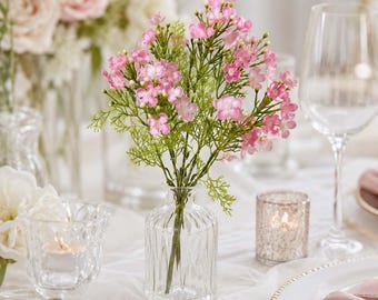 May include: A small bouquet of artificial pink flowers with green stems arranged in a clear, vertically ribbed glass vase. The flowers feature multiple tiny petals and buds, creating a delicate, branched appearance within the transparent container.
