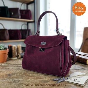 Könnte beinhalten: Eine tiefburgunderrote Wildlederhandtasche mit strukturiertem Tragegriff, einer Umschlagklappe mit silberfarbenem Drehverschluss aus Metall und einem passenden Schulterriemen. Die Tasche steht auf einer Holzoberfläche neben einer kleinen Karte.