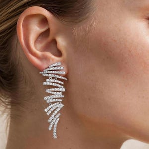 May include: A long, silver-toned earring featuring multiple layered, curved bars encrusted with small, clear, sparkling stones. The design follows a cascading, fan-like shape that extends downwards from the earlobe, creating a textured, linear look.