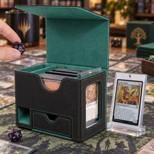 May include: A black and green deck box with a hinged lid, featuring a front-facing display window for a trading card and a small storage drawer. The interior is lined with green fabric, and the exterior is black with green stitching.