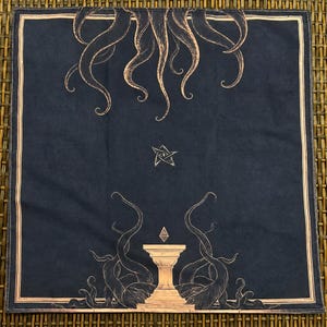 Può includere: Un panno quadrato in tessuto nero con un'illustrazione a linee color marrone chiaro. Il design include tentacoli che scendono dall'alto, un simbolo a stella centrale e un piedistallo con una forma geometrica fluttuante circondata da tentacoli.