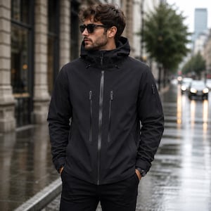 Può includere: Una giacca nera con cappuccio dotata di cerniera frontale integrale, due tasche verticali con zip sul petto e una piccola tasca con zip sulla manica sinistra. La giacca è realizzata in un materiale liscio e resistente all'acqua.