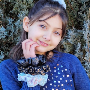 May include: A young girl wearing a blue sweater with silver sequins and a stack of colorful scrunchies on her wrist. The scrunchies are black, gold, and white with a sparkly design.