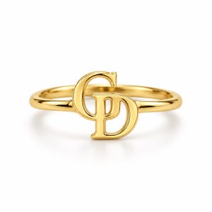 May include: A polished gold ring featuring a central monogram design with the letters C and D intertwined. The band is smooth and circular, showcasing a bright, reflective metallic finish that highlights the elegant, interconnected letter forms.
