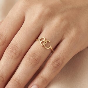 May include: A gold-toned ring featuring two interconnected capital letters, B and C, forming the centrepiece of the band. The letters are rendered in a clean, sans-serif font style, creating a continuous design that sits atop the finger.