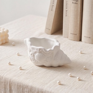 Può includere: Una ciotola in ceramica bianca opaca a forma di conchiglia, caratterizzata da intricati dettagli in rilievo di conchiglie e coralli sulla superficie esterna. La ciotola presenta un bordo smerlato e un interno liscio e cavo, in tinta neutra.