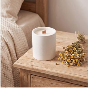 May include: A cylindrical white ceramic candle container with a flat wooden wick centered in white wax. The exterior of the vessel features a subtle, embossed circular floral pattern. Beside the candle lies a small bundle of dried chamomile flowers.