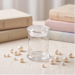 May include: A clear glass cylindrical jar with a matching glass lid, featuring a thick rim and base. The jar is surrounded by numerous small, round, cream-colored faux pearls scattered across the surface, creating a decorative arrangement.
