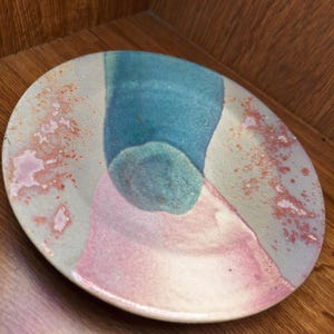 May include: A round ceramic plate featuring a speckled beige base with a prominent vertical blue stripe and a soft pink wash at the bottom. The surface is decorated with scattered orange and rust-coloured spots, creating a textured appearance.