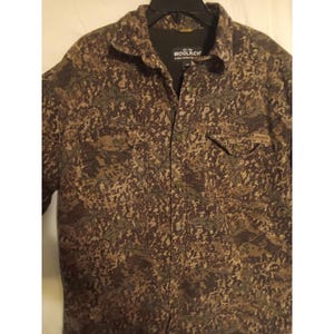 May include: A long-sleeve button-up shirt featuring a brown, black, and tan camouflage pattern. The shirt has a collar, two chest pockets with flaps, and a black interior lining. The Woolrich brand label is visible on the inside neck area.