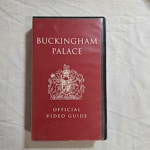 Peut inclure: Un boîtier de cassette VHS rectangulaire avec une couverture rouge foncé. Le texte BUCKINGHAM PALACE est imprimé en blanc en haut, avec OFFICIAL VIDEO GUIDE en bas. Un emblème royal blanc est centré entre le texte sur la couverture.