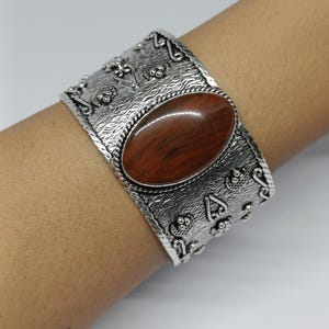 Peut inclure: Un large bracelet manchette de couleur argentée orné d'une grande pierre ovale lisse brun rougeâtre au centre. La surface métallique est fortement texturée avec des motifs en relief complexes, incluant de petits points et des volutes.