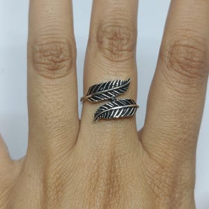 May include: A silver-toned wrap ring featuring two detailed feather motifs that overlap on the finger. The metal has an oxidized finish in the recessed areas to highlight the etched lines and texture of the feathers, creating a distinct contrast.