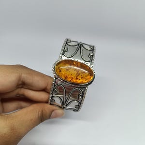 May include: A silver cuff bracelet featuring an oval-shaped amber gemstone at the centre. The metal band displays intricate filigree scrollwork patterns and a textured border, showcasing a polished finish with dark oxidised detailing in the crevices.