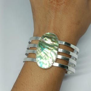 Può includere: Un bracciale a fascia color argento con un grande inserto ovale in conchiglia di abalone. Il bracciale è composto da quattro fasce metalliche parallele collegate al pezzo centrale iridescente, che mostra tonalità vorticose verdi, blu e madreperlacee.