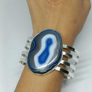 May include: A cuff bracelet featuring a large, polished, oval-shaped blue and white banded agate stone set in a silver-toned metal frame. The band consists of four parallel, thin, curved silver-toned metal strips that wrap around the wrist.