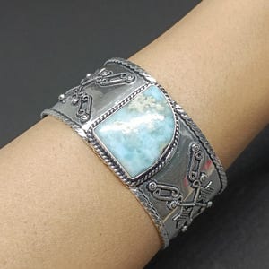 Puede incluir: Una pulsera de brazalete ancha de plata con una piedra central pulida de color azul claro y blanco veteado. La banda de metal está decorada con intrincados patrones geométricos en relieve y bordes trenzados en los extremos superior e inferior.