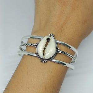 Puede incluir: Una pulsera de plata con una concha de cauri blanca central engastada en un marco de metal decorativo. La pulsera consta de tres bandas distintas, con la banda central que presenta una textura de cuerda trenzada y las bandas exteriores lisas.