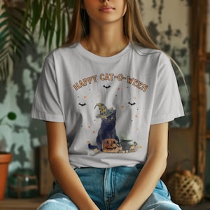 Puede incluir: Una camiseta blanca de manga corta con un gráfico de un gato negro con sombrero de bruja, sentado junto a una calabaza tallada y un caldero. El texto "HAPPY CAT-O-WEEN" aparece impreso sobre el gato, junto a pequeñas ilustraciones de murciélagos.