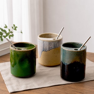 May include: Three cylindrical ceramic cups with a built-in slot on the rim for a metal straw. The left cup is solid green, the center cup is cream with a blue drip glaze, and the right cup features a dark brown base transitioning to a teal top.