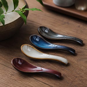 Może przedstawiać: Zestaw czterech ceramicznych łyżek do zupy ułożonych w rzędzie, każda z wyraźną błyszczącą glazurą w kolorze głębokiej czerwieni, kremowym, kobaltowym i antracytowym. Łyżki mają zakrzywiony uchwyt oraz gładkie, odblaskowe wykończenie powierzchni.